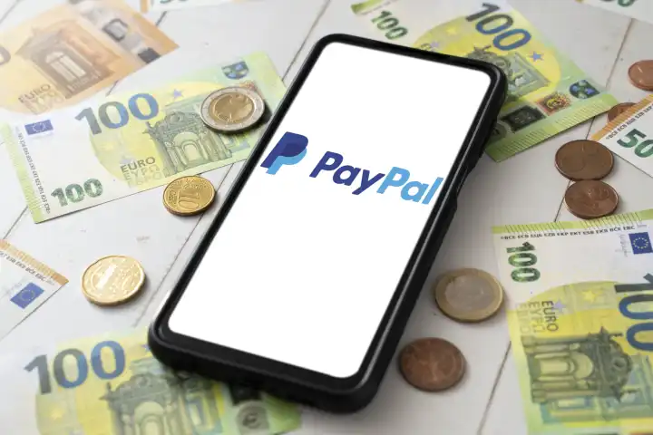 8 August 2023: Smartphone with Paypal logo on screen next to euro bills and coins PHOTOMONTAGE