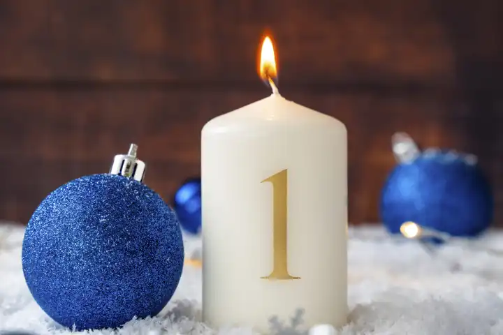 Erster Advent, brennende Kerze in Schnee mit blauen Christbaumkugeln. Kerze mit der Zahl 1