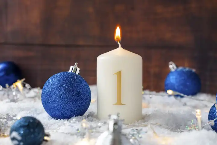 11 August 2023: First Advent, burning candle in snow with blue Christmas tree balls. Candle with the number 1