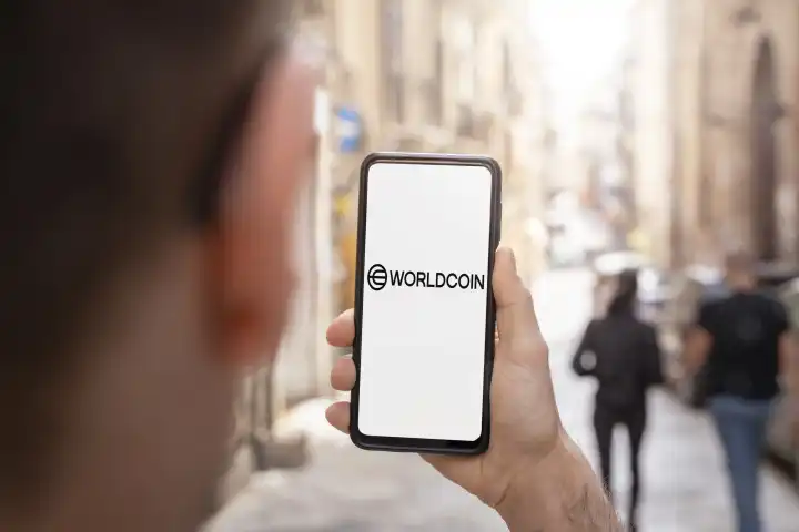 Mann hält Smartphone in der Hand mit  Worldcoin Logo auf dem Bildschirm. Symbolbild neue Kryptowährung Worldcoin. Für den Worldcoin ist eine World ID mit Iris Augen Scan notwendig FOTOMONTAGE