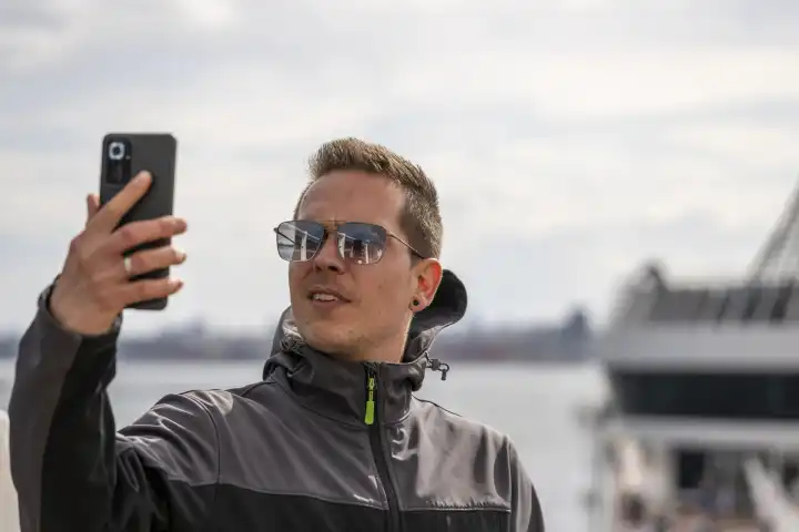 Mann hält ein Smartphone auf einem Kreuzfahrtschiff. Internet und Mobilfunk Konzept. Urlauber mit Sonnenbrille und Jacke auf einem Schiff