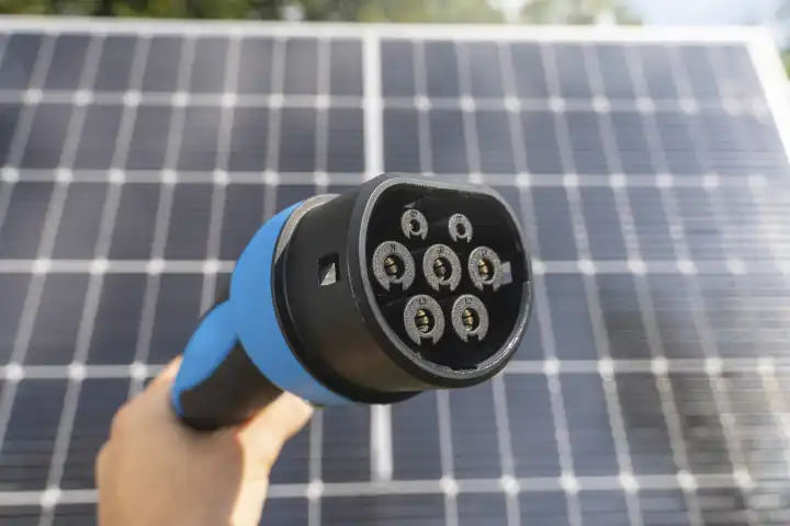 15 August 2023: Hand with charging plug for electric vehicles in front of a solar installation. Photovoltaic energy for charging an electric car concept. Green energy for mobility