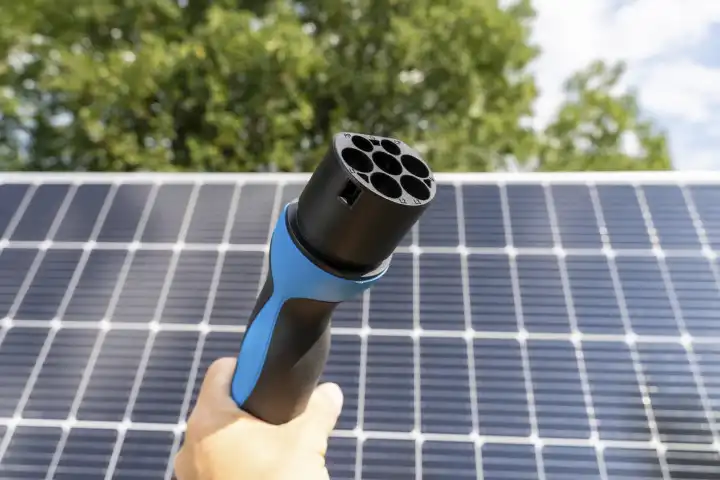 15 August 2023: Hand with charging plug for electric vehicles in front of a solar installation. Photovoltaic energy for charging an electric car concept. Green energy for mobility