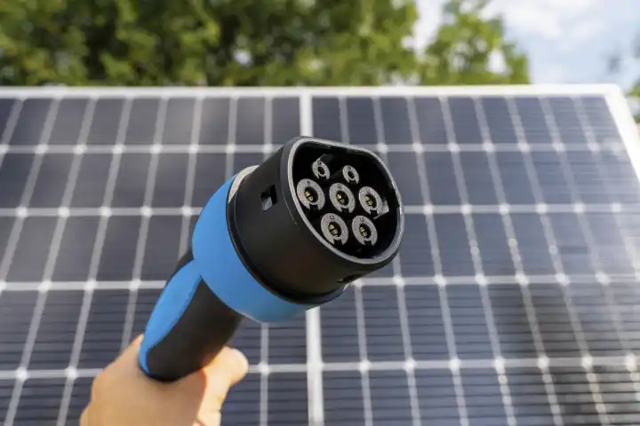Hand mit Ladestecker für Elektrofahrzeuge vor einer Solaranlage. Photovoltaik Energie für die Aufladung von einem Elektro Auto Konzept. Grüne Energie für Mobilität