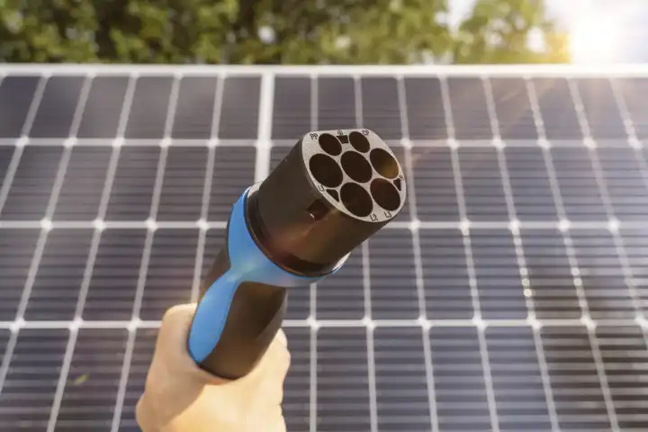 Hand mit Ladestecker für Elektrofahrzeuge vor einer Solaranlage. Photovoltaik Energie für die Aufladung von einem Elektro Auto Konzept. Grüne Energie für Mobilität