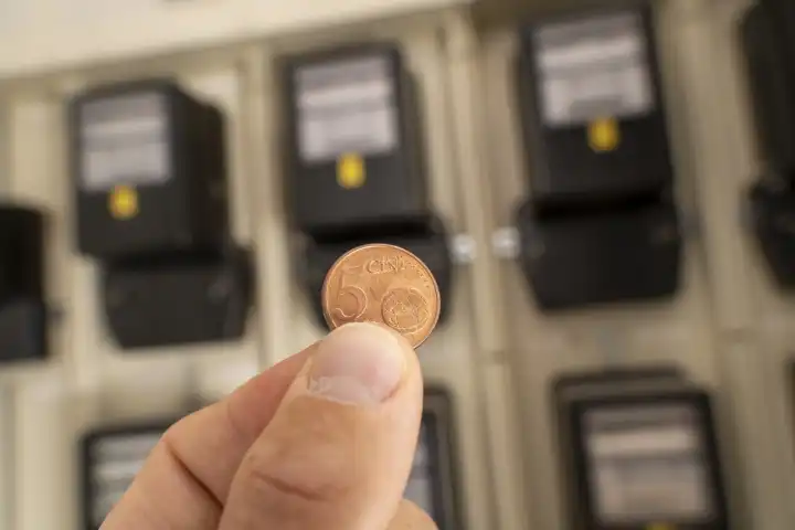 Bavaria, Germany - 25 August 2023: Hand holding a 5 euro cent coin in front of electricity meter. Symbol image industrial electricity price