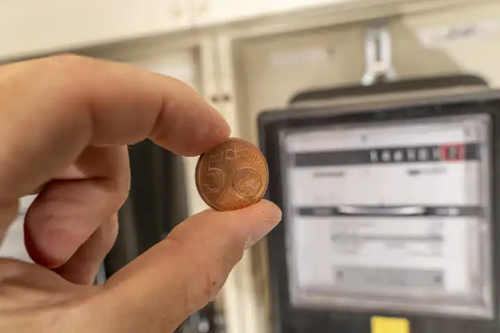 Hand hält eine 5 Euro Cent Münze vor Stromzähler. Symbolbild Industriestrompreis