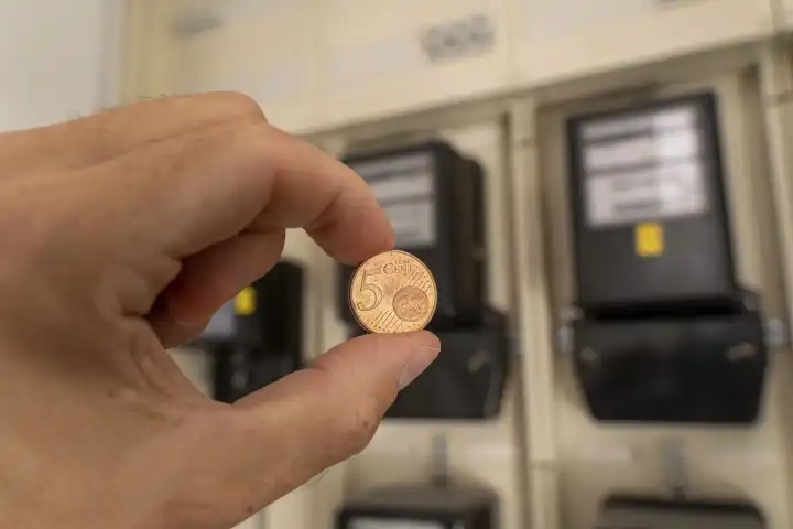 Bavaria, Germany - 25 August 2023: Hand holding a 5 euro cent coin in front of electricity meter. Symbol image industrial electricity price