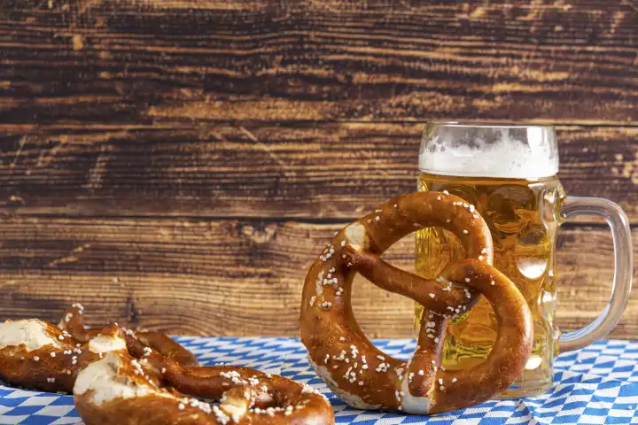 Bavaria, Germany - 25 August 2023: Oktoberfest background, Bavarian flag with beer in Maßkrug and fresh brown pretzels. Wooden background with text free space