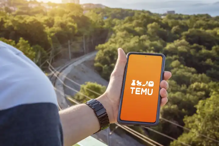26 August 2023: TEMU Online Marketplace app on the screen of a smartphone. Man holding cell phone with Temu logo in hand PHOTOMONTAGE