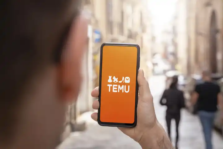 TEMU Online Marktplatz App auf dem Bildschirm von einem Smartphone. Mann hält Handy mit Temu Logo in der Hand FOTOMONTAGE