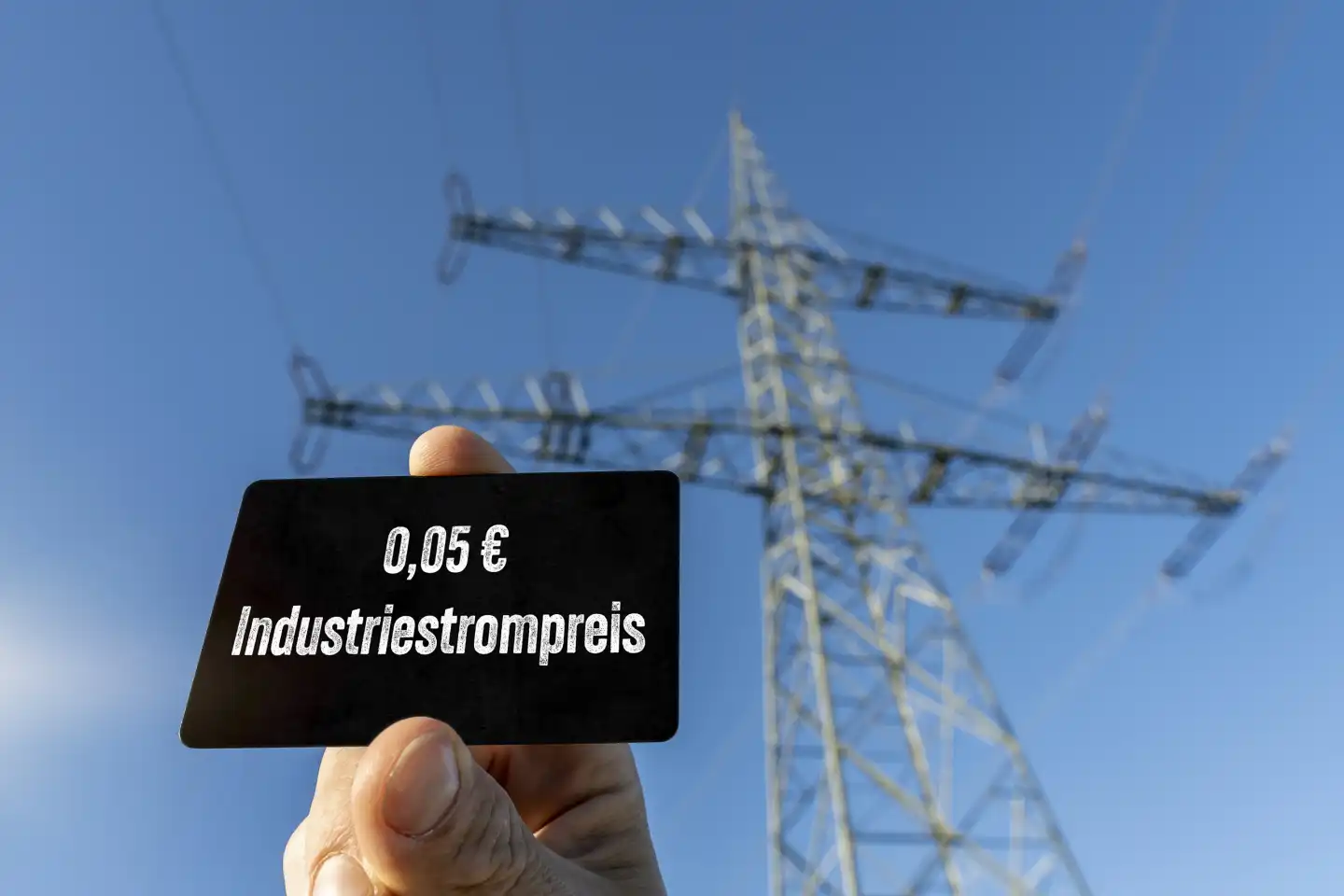 Augsburg, Bavaria, Germany - August 7, 2023: Hand holding plaque in front of a high voltage power pole with the inscription: Industrial electricity price 5 cents PHOTOMONTAGE