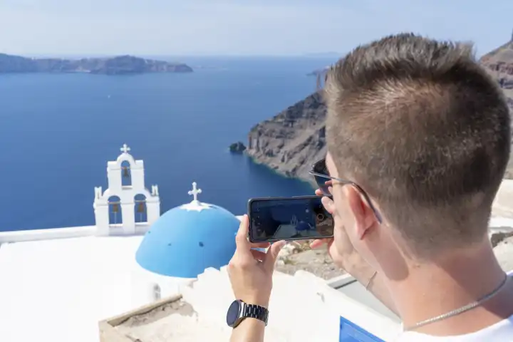 Ein Urlauber macht Fotos von einer Kapelle mit einer blauen Kuppel auf Santorini in Griechenland im Sommer mit seinem Smartphone
