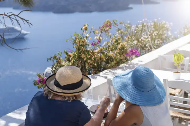 Santorini, Greece - 20 September 2023: Two ladies in a restaurant on Santorini in Greece during nice weather. Vacation and travel concept. Women in sun hats on white chairs eating with sea view
