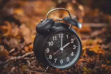 Augsburg, Bavaria, Germany - 7 October 2023: Time change symbol image, black alarm clock between autumn leaves. Change from summer time to winter time