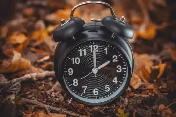 Augsburg, Bavaria, Germany - 7 October 2023: Time change symbol image, black alarm clock between autumn leaves. Change from summer time to winter time