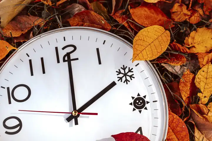 Augsburg, Bavaria, Germany - 16 October 2023: Symbolic image time change from summer time to winter time. Clock lies between foliage in autumn with the hand on time 2 o'clock. Sun and ice symbol as time PHOTOMONTAGE