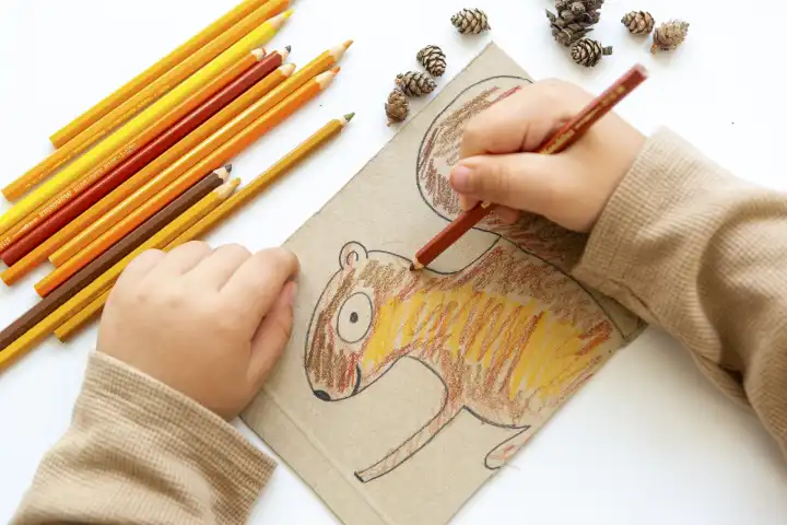 Kinderspaß mit Herbstlaub: Kreatives Basteln zu Hause. Ein Kind bastelt ein Eichhörnchen das es selbst gemalt hat und mit Tannenzapfen dekoriert. Hände malen mit Farbe das Bild aus