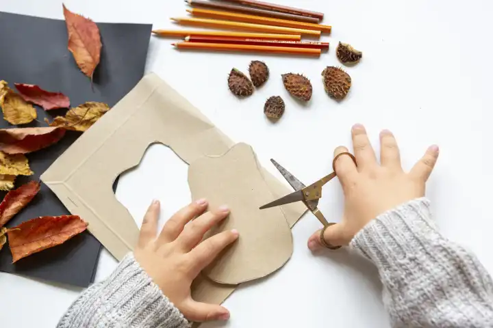 Kinderspaß mit Herbstlaub: Kreatives Basteln zu Hause. Ein Kind bastelt einen Igel aus Pappe. Hände schneiden die Form aus einem Pappkarton mit der Schere aus