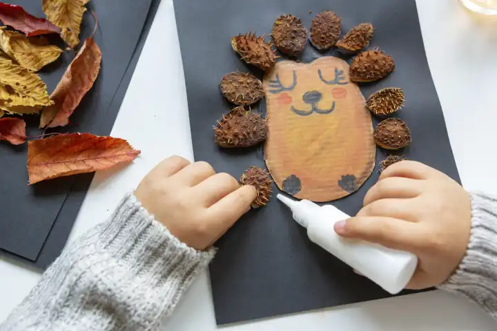 Kinderspaß mit Herbstlaub: Kreatives Basteln zu Hause. Ein Kind bastelt eine Kollage von einem Igel. Hände kleben Laub und Kastanien Schalen aus das Bild