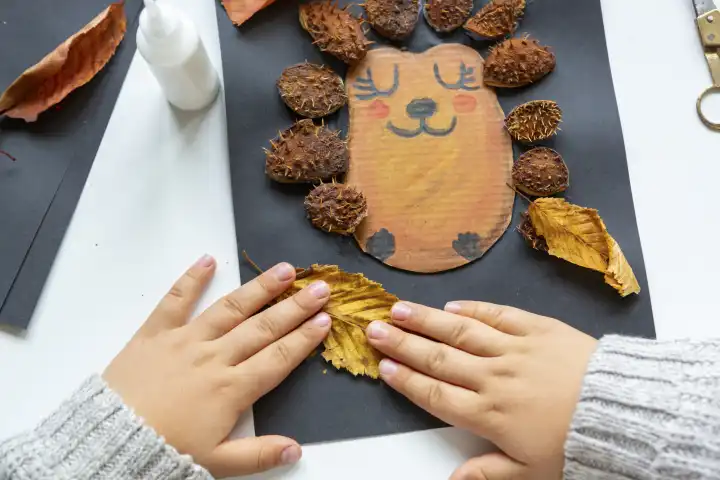 Kinderspaß mit Herbstlaub: Kreatives Basteln zu Hause. Ein Kind bastelt eine Kollage von einem Igel. Hände kleben Laub und Kastanien Schalen aus das Bild