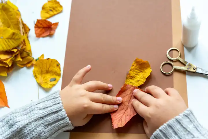 Kinderspaß mit Herbstlaub: Kreatives Basteln zu Hause. Ein Kind bastelt mit leerem Papier und Karton und bunten Blättern im Herbst