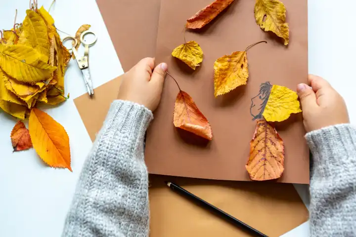 Kinderspaß mit Herbstlaub: Kreatives Basteln zu Hause. Ein Kind bastelt mit leerem Papier und Karton und bunten Blättern im Herbst