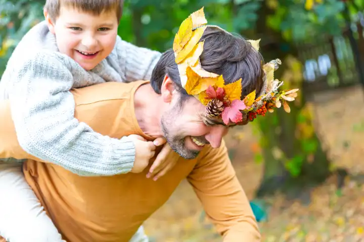 Papa und Sohn spielen im Herbst im Garten. Kleiner Junge Huckepack auf dem Rücken von seinem Vater mit einer herbstlichen selbstgebastelten Krone aus bunten Blättern. Familie im Herbst Konzept