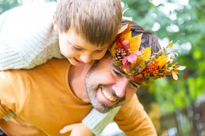 Papa und Sohn spielen im Herbst im Garten. Kleiner Junge Huckepack auf dem Rücken von seinem Vater mit einer herbstlichen selbstgebastelten Krone aus bunten Blättern. Familie im Herbst Konzept