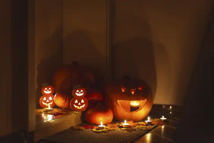 Halloween Dekoration vor einer Wohnungstüre bei Dunkelheit. Brennende Kerzen leuchten um gruselige verzierte Halloween Kürbisse vor einer Türe