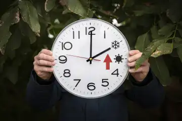 Mann hält große runde Uhr mit Sommer und Winterzeit in der Hand. Ein großer roter Pfeil steht für die Uhr zurückstellen FOTOMONTAGE