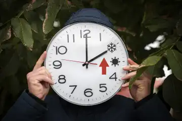 Mann hält große runde Uhr vor sein Gesicht mit Sommer und Winterzeit in der Hand. Ein großer roter Pfeil steht für die Uhr zurückstellen. Uhrzeit umstellen erinnerung FOTOMONTAGE
