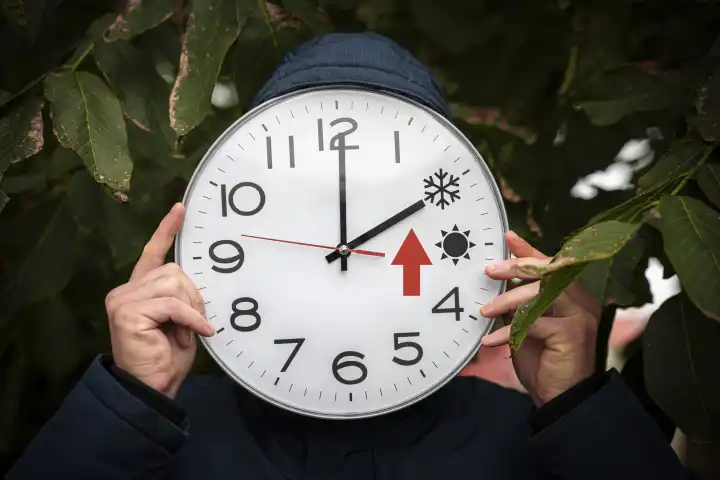 24 October 2023: Man holding big round clock in front of his face with summer and winter time in hand. A big red arrow stands for clock reset. Change time reminder PHOTOMONTAGE