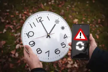 Mann hält große runde Uhr mit Winterzeit und Sommerzeit in der Hand. Daneben ein Smartphone mit einem Warnsymbol. Digitale Umstellung der Uhrzeit und Funkuhr Konzept FOTOMONTAGE