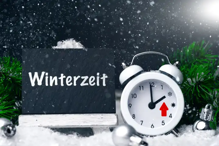27 October 2023: Panel with text winter time next to a white alarm clock during snowfall in winter environment. Sign next to clock. Time change from summer time to winter time PHOTOMONTAGE