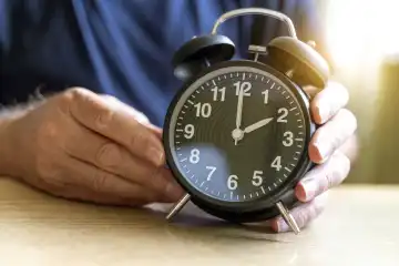 Zeitumstellung auf Winterzeit, Ein alter Mann hält einen Wecker bei dem er die Uhr umgestellt hat von Sommerzeit auf Winterzeit