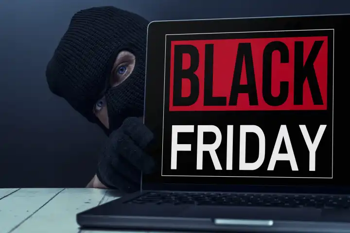 Laptop Computer mit Black Friday Schriftzug auf dem Bildschirm und einem maskierten Verbrecher neben dem Computer, Symbolbild Betrug und Abzocke zum Black-Friday FOTOMONTAGE