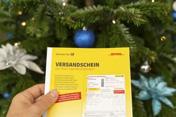 Augsburg, Bavaria, Germany - December 15, 2023: DHL parcel and parcel shipping label is held by a man in front of a decorated Christmas tree. Sending gifts with a parcel concept