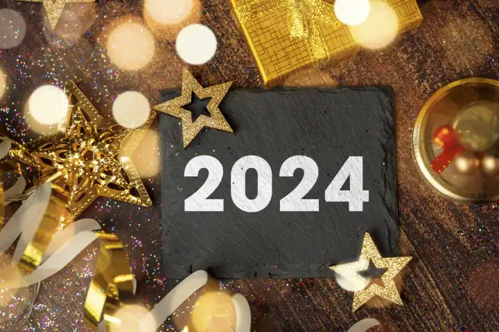 Symbolbild Neujahr Silvester 2024. Schiefertafel umgeben von goldener Dekoration mit der Aufschrift 2024 FOTOMONTAGE