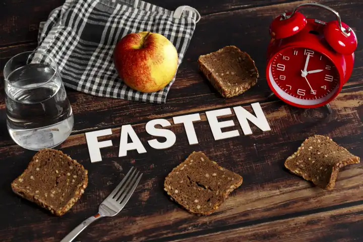 January 31, 2024: Symbolic image of intermittent fasting and fasting, The writing train fasting surrounded by dry bread, apple and a glass of water as well as a red alarm clock