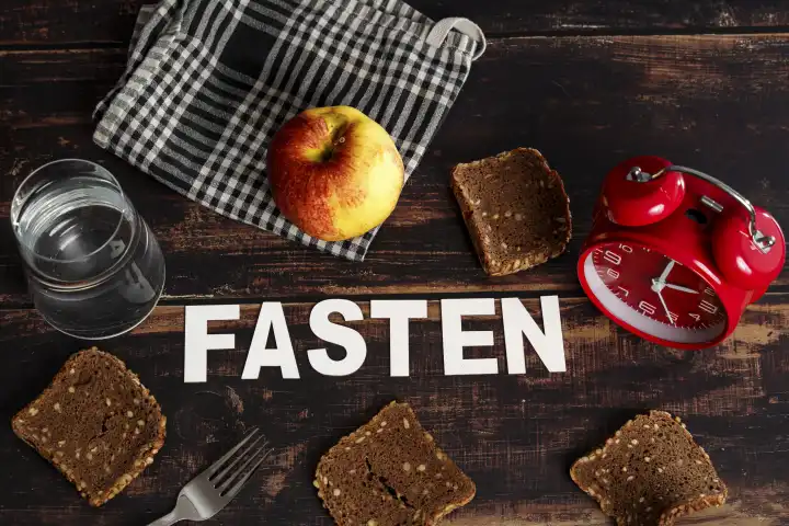 Symbolbild Intervallfasten und Fasten, Der Schrifzug Fasten umgeben von trockenem Brot, Apfel und einem Glas Wasser sowie einem roten Wecker