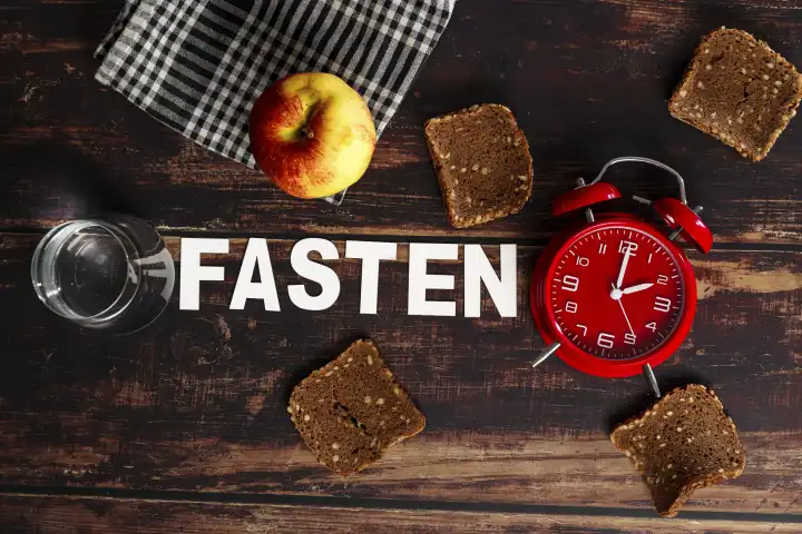 January 31, 2024: Symbolic image of intermittent fasting and fasting, The writing train fasting surrounded by dry bread, apple and a glass of water as well as a red alarm clock