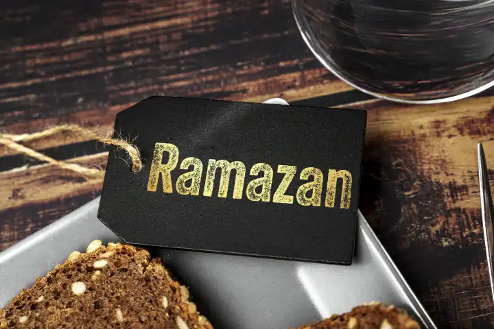 Ramazan (Türkisch für Ramadan), der Fastenmonat der Muslime. Schriftzug auf einer Tafel auf einem Teller mit einem Stück Brot neben einem Glas Wasser FOTOMONTAGE