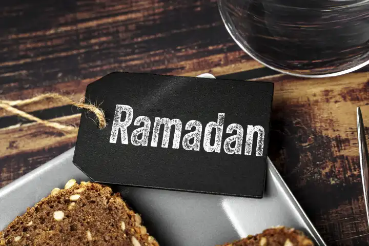 Ramadan, der Fastenmonat der Muslime. Schriftzug auf einer Tafel auf einem Teller mit einem Stück Brot neben einem Glas Wasser FOTOMONTAGE