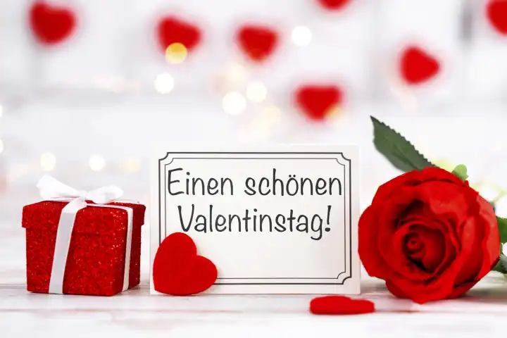 Einen schönen Valentinstag! Gruß auf einer Grußkarte neben einem Geschenk und einer roten Rose FOTOMONTAGE