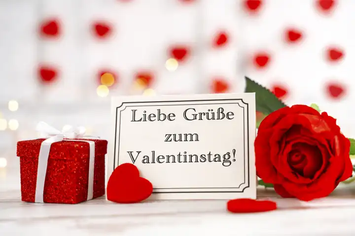 Liebe Grüße zum Valentinstag! Gruß auf einer Grußkarte neben einem Geschenk und einer roten Rose FOTOMONTAGE
