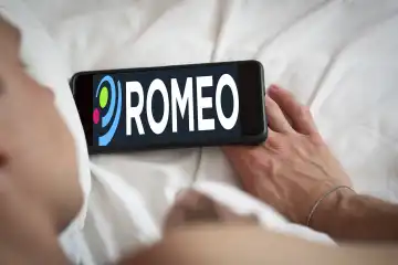 Ein Mann liegt auf dem Bett mit seinem Handy. Auf dem Handy steht das Logo der App für schwule Männer namens Romeo, ehemals Gayromeo. FOTOMONTAGE