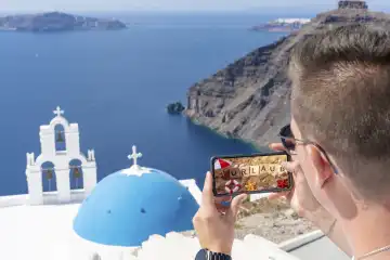 Symbolbild Urlaub, Junger Mann im Urlaub, macht ein Foto mit seinem Smartphone in Griechenland, vor dem blauen Meer und schöner Landschaft. Auf dem Handy ein Bild vom Strand mit dem Text: URLAUB. FOTOMONTAGE