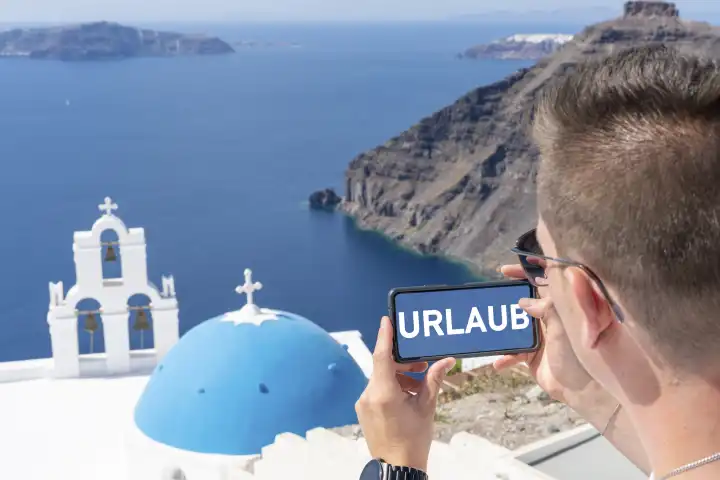 February 11, 2024: Symbolic picture vacation, young man on vacation, takes a photo with his smartphone in Greece, in front of the blue sea and beautiful landscape. On the phone a picture in colors of the water with the text: HOLIDAY. PHOTOMONTAGE