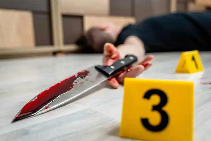 Symbolbild Tatort eines Mordfalls (gestellte Szene). Ein Mann liegt tot auf dem Boden in einer Wohnung. Markierungen von Beweisstücken der Polizei und mit Blut verschmierte Tatwaffe, ein Messer